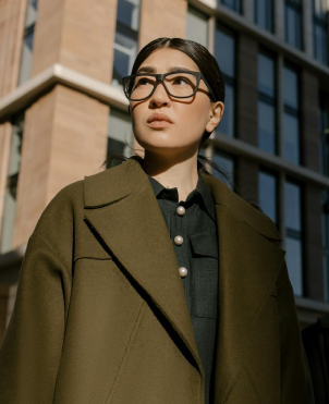 Confident woman wearing black-framed glasses and an olive green coat standing outdoors in front of a modern building.