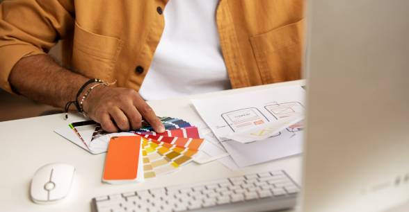 Person in orange shirt selecting colors from a color swatch book on a desk with design sketches and a computer keyboard and mouse.